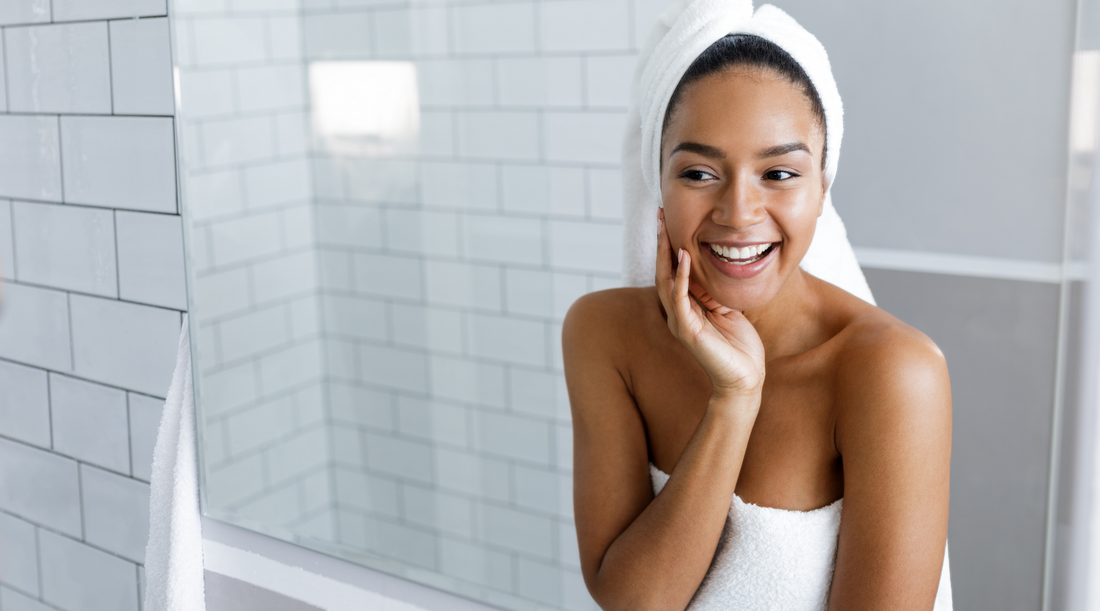 Woman with glowing skin looking in bathroom mirror. Learn how to pick the right serums for your skin concerns 